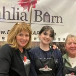 Three smiling women posing at a Dahlia Barn booth; two wear aprons with the Dahlia Barn logo, banner in the background.