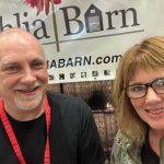 Two smiling adults pose in front of a 'Dahlia Barn' banner at an event or booth.