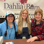 Three women posing at a booth in front of a 'Dahlia Barn' banner, smiling for the camera.